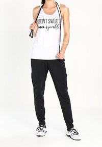 Débardeur blanc avec texte noir "NE TRANPIRE PAS, ÉTINCELLE" associé à un pantalon de jogging noir et des chaussures de sport. Le mannequin tient une corde à sauter.