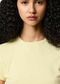 Jasnożółta bawełniana koszulka z okrągłym dekoltem i krótkimi rękawami, prezentująca gładką teksturę na tle kręconych, ciemnych włosów.