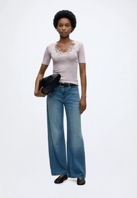 Γυναίκα με κοντά κατσαρά μαλλιά που φοράει ανοιχτό ροζ τοπ με κεντητά λουλούδια, φαρδιά μπλε τζιν, μαύρη ζώνη, μαύρα παπούτσια και κρατάει μαύρη διπλωμένη τσάντα.