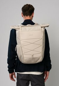 Sac à dos beige à rabat, en tissu texturé, avec un détail de laçage à l'avant, une poche latérale et des bretelles réglables, présenté sur un fond neutre.