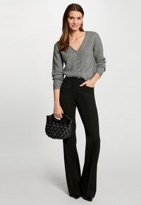 Grijze geribbelde cardigan met een V-hals en knoopsluiting, gecombineerd met zwarte flare broeken. Model houdt een zwarte gewatteerde handtas vast.