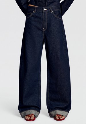 Person iført mørkeblå vidde jeans med optrukne opkanter og røde åbne sandaler, der viser malede tånegle.