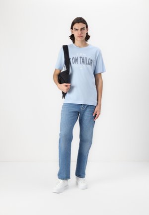 Lyseblå T-shirt med "TOM TAILOR" grafik, parret med lyseblå jeans og hvide sneakers. Sort skuldertaske bæres på venstre skulder.