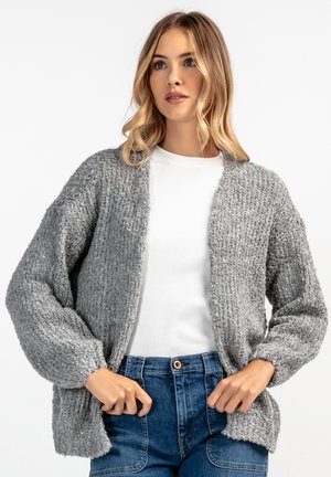 Szary sweter z dzianiny o teksturowej powierzchni, z otwartym przodem i opadłymi ramionami, zestawiony z białym t-shirtem i niebieskimi dżinsami z wysokim stanem.