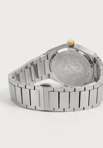 Montre bracelet en acier inoxydable argenté avec tête de Méduse gravée au dos, couronne dorée et "Versace" gravé sur la boucle métallique du bracelet.