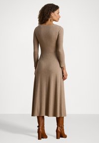 Donna con capelli ricci che indossa un abito midi beige a maniche lunghe e stivali con tacco marroni, in piedi e girata di spalle contro uno sfondo neutro.