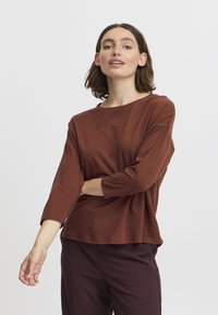 Maglietta marrone a maniche lunghe, morbida e dal taglio ampio, con scollatura rotonda. Tessuto morbido, leggermente texture, abbinata a pantaloni di un borgogna scuro.
