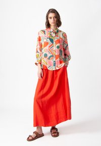 Veelkleurige blouse met geometrische patronen, gecombineerd met een lange, vlotte rode linnen rok en bruine sandalen.