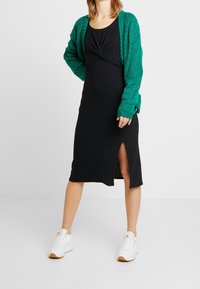Svart midiklänning med ribbat mönster och knut framtill, i kombination med en grön stickad cardigan med texturerat kabelmönster. Vita sneakers.
