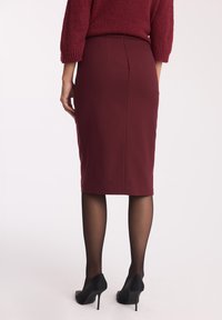 Bordowa spódnica ołówkowa o gładkiej teksturze z tylnym plisowaniem, w połączeniu z przezroczystymi czarnymi rajstopami i czarnymi butami na obcasie.