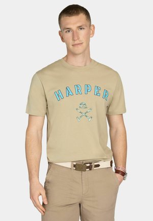 Joven con camiseta beige "HARPER" con logo de remos cruzados, pantalones beige, cinturón blanco, reloj y pulseras, de pie con una mano en el bolsillo.