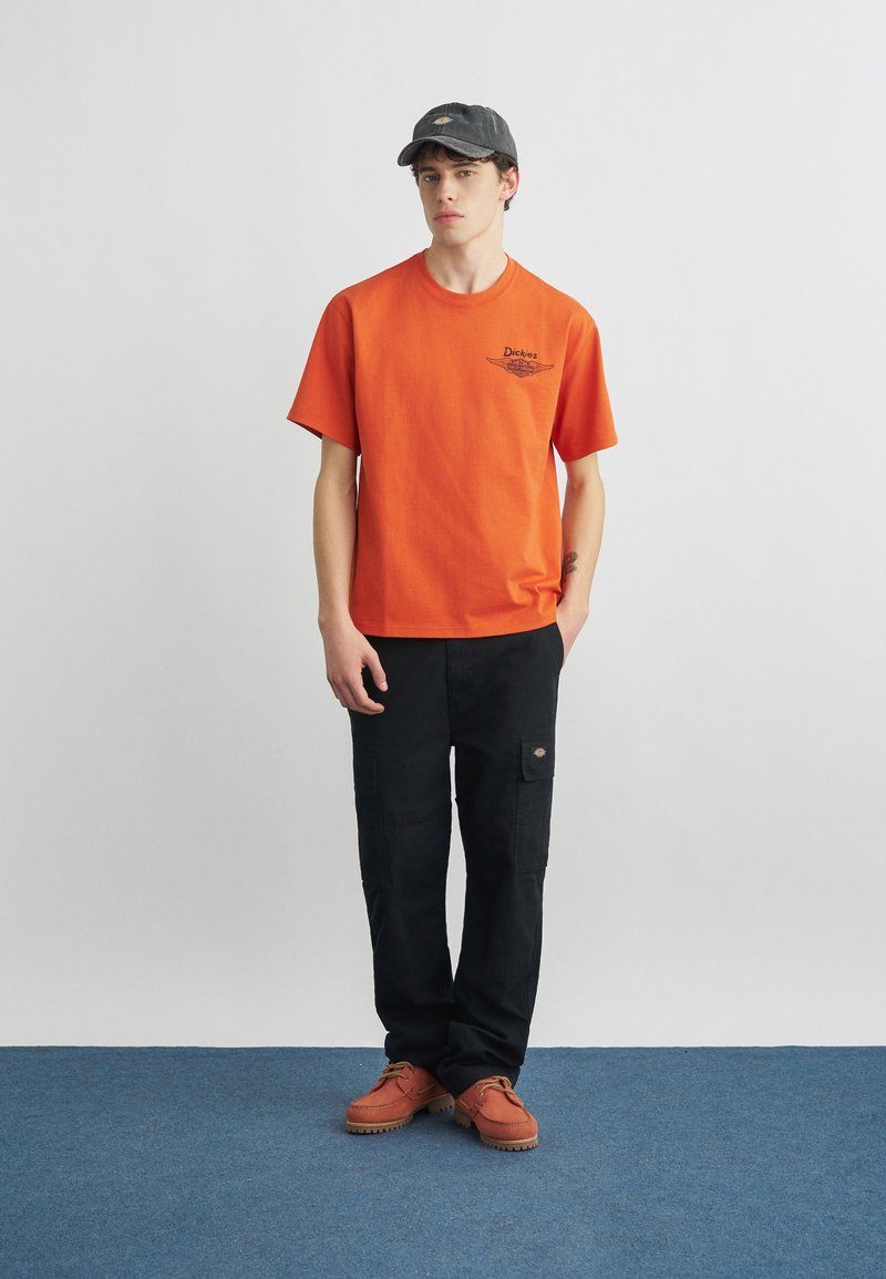 Junger Mann, der ein oranges Dickies-T-Shirt, schwarze Cargohosen, orangefarbene Schuhe und eine schwarze Dickies-Baseballkappe trägt und auf einem blauen Teppich steht.