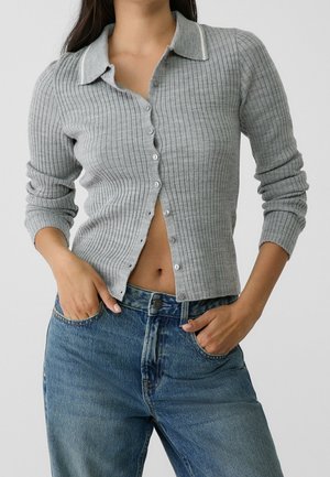 Frau trägt einen taillierten grauen, gerippten Strickpullover mit Knöpfen und Kragen sowie blaue hoch taillierte Jeans, Hände in den Taschen, vor einem schlichten Hintergrund.