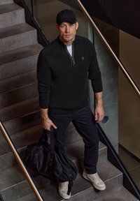 Sudadera negra con cremallera y tejido texturizado, pantalones negros y zapatillas beige. Lleva una bolsa de deporte negra, de pie en escaleras de concreto.