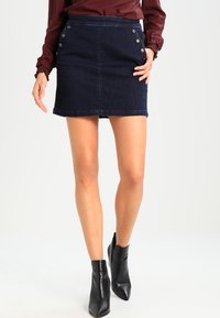 Donna che indossa una mini gonna in denim blu scuro con bottoni laterali, un top a maniche lunghe bordeaux e stivaletti neri alla caviglia, in piedi su uno sfondo bianco.