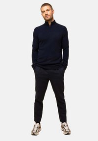 Granatowy sweter na zamek z dzianiny, zestawiony z ciemnymi spodniami i białymi sneakersami. Model stoi z rękami w kieszeniach na neutralnym tle.