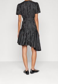 Robe noire avec des manches bouffantes courtes, en tissu texturé, taille ceinturée, ourlet asymétrique évasé, associée à des ballerines noires.