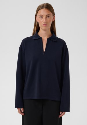 Femme aux cheveux bruns et raides portant un pull bleu marine à manches longues avec un col et une courte fermeture éclair à l'avant, ainsi qu'un pantalon noir.