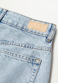 Jean en denim bleu clair en gros plan montrant la poche arrière, la boucle de ceinture et une étiquette en cuir beige avec le texte "MNG DNM 1984" sur fond blanc.