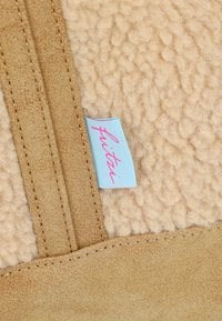 Suède synthétique beige avec une surface texturée douce. Présente une étiquette bleu clair avec un texte rose. Comprend des coutures le long des bords.