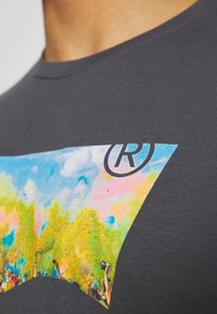 Primer plano de una camiseta gris oscura con un gráfico colorido de una multitud lanzando polvo de colores, que incluye un símbolo de marca registrada.