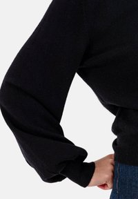 Pull en maille noire avec manches longues bouffantes et taille ajustée, doté d'une texture douce et de poignets côtelés, associé à un jean denim bleu.