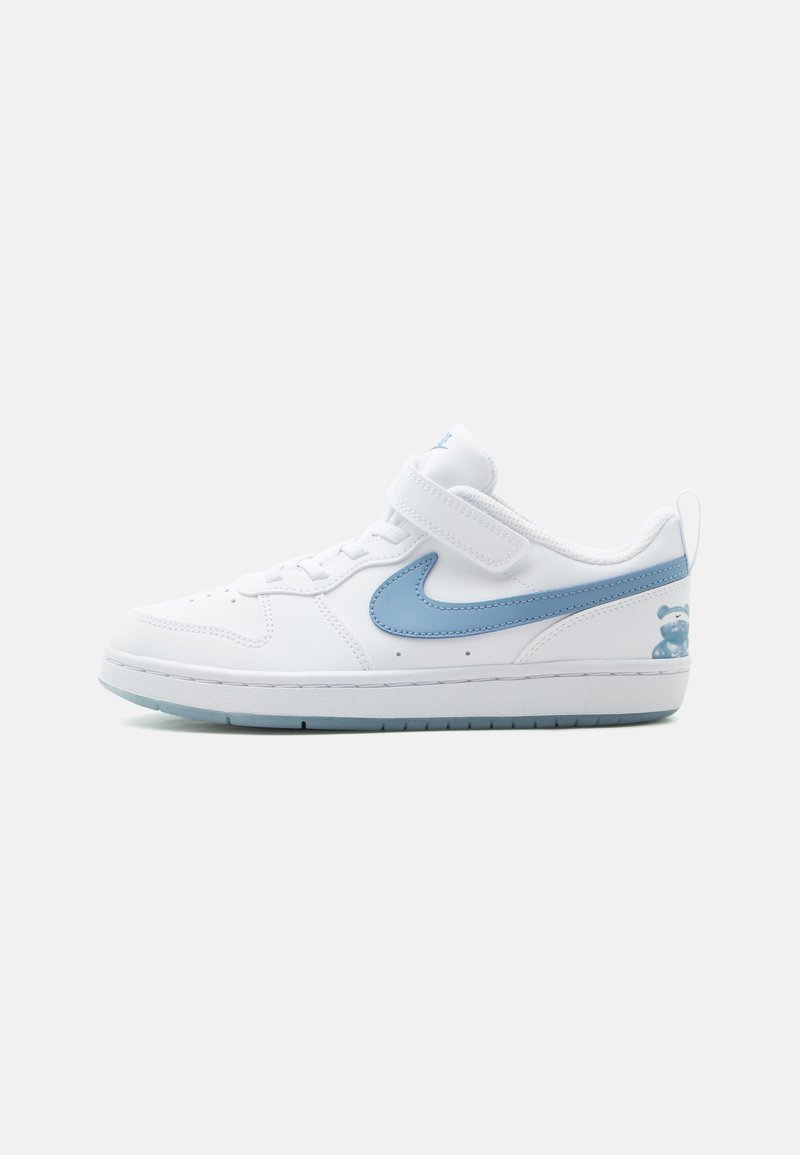 Άσπρο sneaker χαμηλού τύπου με γαλάζιο λογότυπο Nike, ιμάντα με βέλκρο και μικρό γραφικό αρκουδάκι στη φτέρνα, τοποθετημένο σε απλό φόντο.