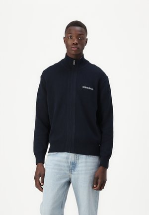 Navy zip-up sweater with high collar and ribbed cuffs. Features "GUESS JEANS" logo on the left chest, made from soft knitted fabric.