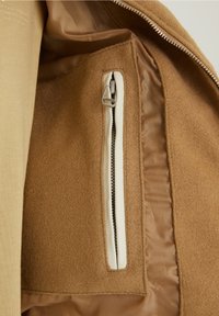 Gros plan sur la poche d'une veste en laine marron avec une fermeture éclair verticale bordée de blanc et une doublure en tissu beige à l'intérieur de la veste.