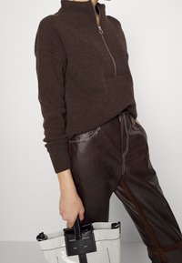 Personne portant un pull à fermeture éclair marron foncé et un pantalon en cuir marron foncé, tenant un sac fourre-tout blanc et noir avec texte de marque visible.