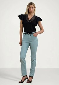 Γυναίκα που φοράει μια μαύρη μπλούζα από δαντέλα με V λαιμόκοψη και φουσκωτά μανίκια, ανοιχτό μπλε ψηλόμεσα τζιν, μαύρη ζώνη και μαύρες πέδιλες με τακούνι.