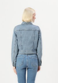 Chaqueta de mezclilla en azul claro con un diseño corto, que presenta un cuello, puños con botón y costuras visibles. Combinada con jeans a juego.