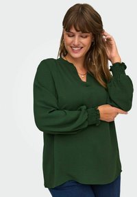 Donna che sorride e aggiusta le maniche lunghe di una blusa verde scuro con polsini arricciati, indossando orecchini a cerchio e una collana con un pendente.