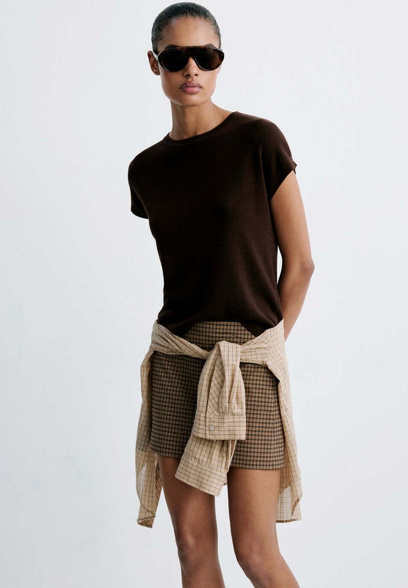 Femme portant un haut à manches courtes marron foncé, une mini-jupe à carreaux, une chemise à carreaux beige nouée à la taille, et de grandes lunettes de soleil foncées sur un fond uni.