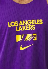 Lila hálós kosárlabda mez, rajta a "LOS ANGELES LAKERS" felirattal és egy sárga logóval. Lélegző anyagból készült és szabványos kerek nyakkivágással rendelkezik.