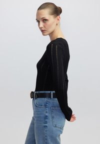 Schwarzes Langarmshirt mit vertikal gerippter Textur, kombiniert mit hochgeschnittenen hellblauen Jeans und einem schwarzen Gürtel mit goldener Schnalle.