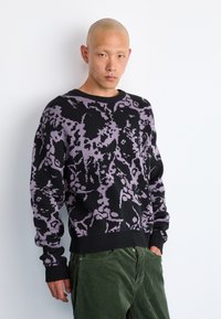 Pulover tricotat în negru cu un model abstract violet, având un guler rotund și manșete ribbate. Asortat cu pantaloni din corduroy verde închis.