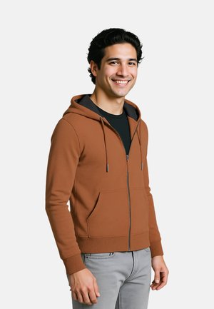 Felpa zip con cappuccio marrone, con coulisse, tasche frontali e polsini a costine. Indossata sopra una maglietta nera, completata da pantaloni grigi.