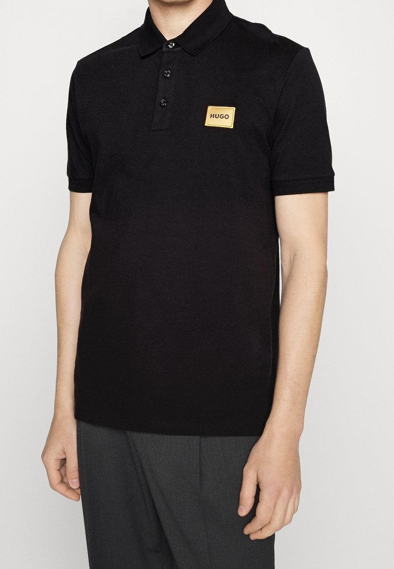Homme portant un polo noir à manches courtes avec un patch doré "HUGO" sur la poitrine et un pantalon gris foncé, devant un fond uni.