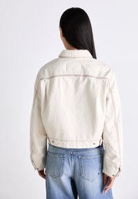Gekraagde cropped jack in off-white canvas met contrasterende stiksels; gecombineerd met lichtblauwe jeans met achterzakken.