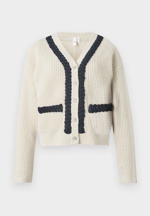 Cardigan en tricot crème avec bordures en tresse noire le long du col en V, de la patte de boutonnage et des poches, avec des manches longues et des boutons en argent.