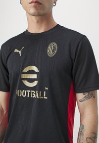 Black football jersey with textured design, gold logos and accents, red side panels, round neckline, and short sleeves.