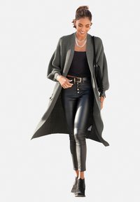 Lange groene open cardigan, ribbelstructuur, gedragen over een zwart topje, gecombineerd met glanzende zwarte leren broek en enkellaarsjes. Zilveren ketting als accent.