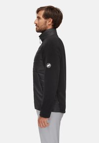 Hombre vistiendo una chaqueta híbrida de acolchado negro y forro polar, de pie en perfil contra un fondo claro y liso.