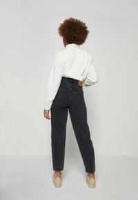 Jeans negros de tiro alto con una cinturilla con bucles, bolsillos traseros y un parche de marca de cuero. Combinados con una camisa blanca de botones y zapatos beige.