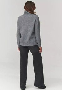 Kobieta stojąca tyłem, ubrana w szary prążkowany sweter i czarne spodnie z szerokimi nogawkami, oparta o jednolitą jasnoszara ścianę.