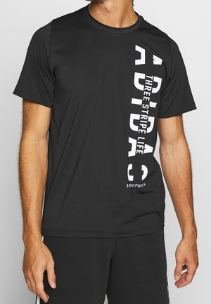 Mężczyzna ubrany w czarną koszulkę Adidas z pionowym białym napisem "ADIDAS" oraz "THREE STRIPE LIFE" po prawej stronie.