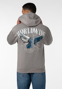 Graue Kapuzenjacke mit einem großen Adler-Graphic auf der Rückseite, mit dem Text "WORLDWIDE" darüber und Tourdetails darunter in sanften Farben.