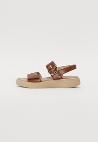 Brown leather sandal with adjustable strap, round gold accent, and a light beige platform sole. Flat footbed with textured base.