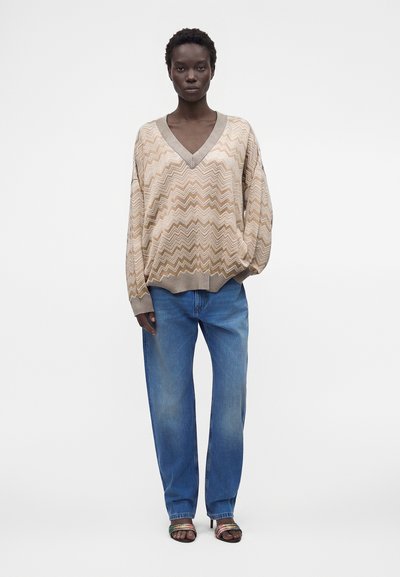 Femme portant un pull beige à motif zigzag, un jean bleu et des sandales ouvertes, debout devant un fond blanc uni.